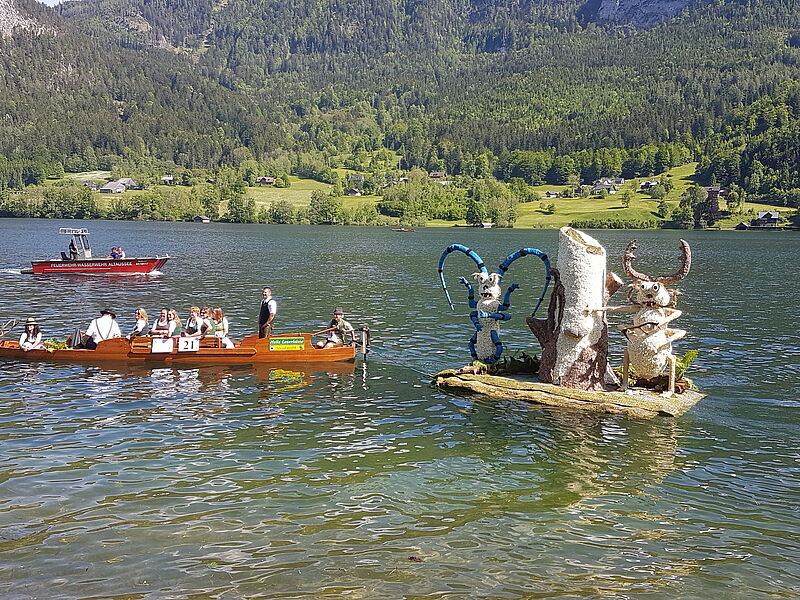 Die Figur wird über den See gezogen von eienr Plätte, in der mehrere menschen mit tracht sitzen.