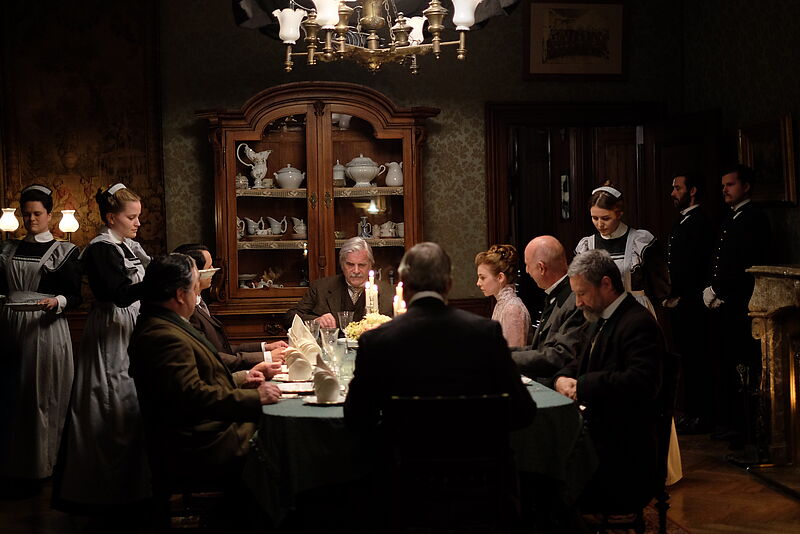 Am Tisch sitzt eine Runde von Menschen beim Essen, im Hintergrund Bedienstete. In der Mitte sitzt Schauspieler Peter Simonischek.