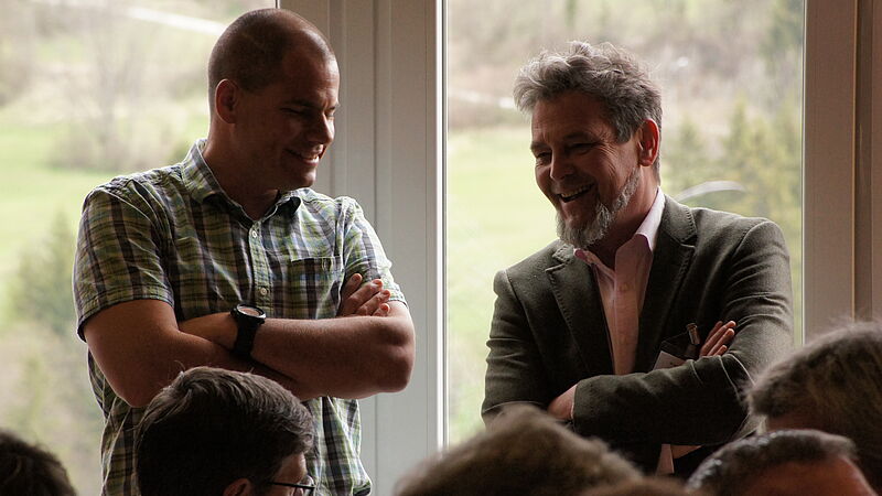 Zwei Männer stehen beinander und unterhalten sich. Beide haben die Arme verkreuzt und schauen freundlich.