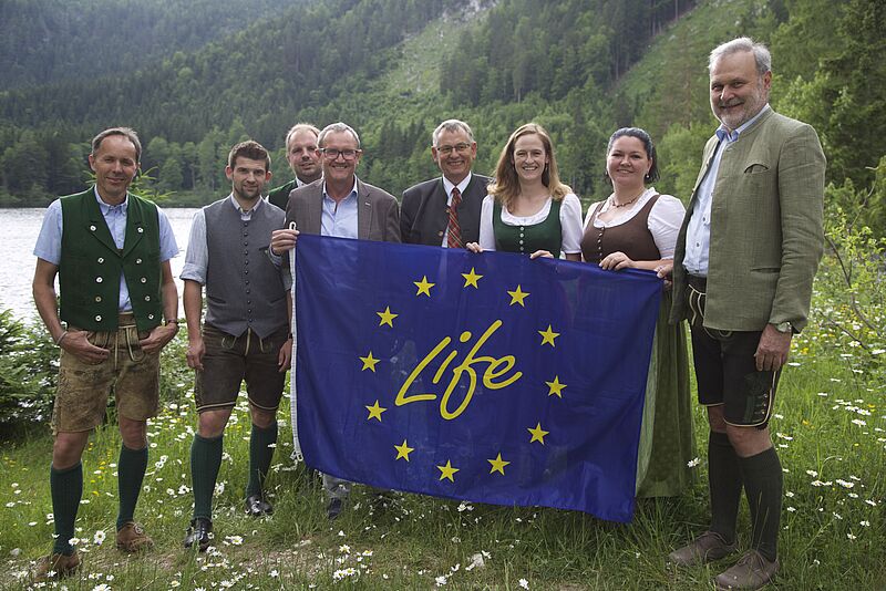 Sechs Männer stehen gemeinsam mit zwei Frauen nebeneinander und sind gekleidet in Tracht. Sie halten eine blaue Fahne auf der LIFE in gelb steht. 