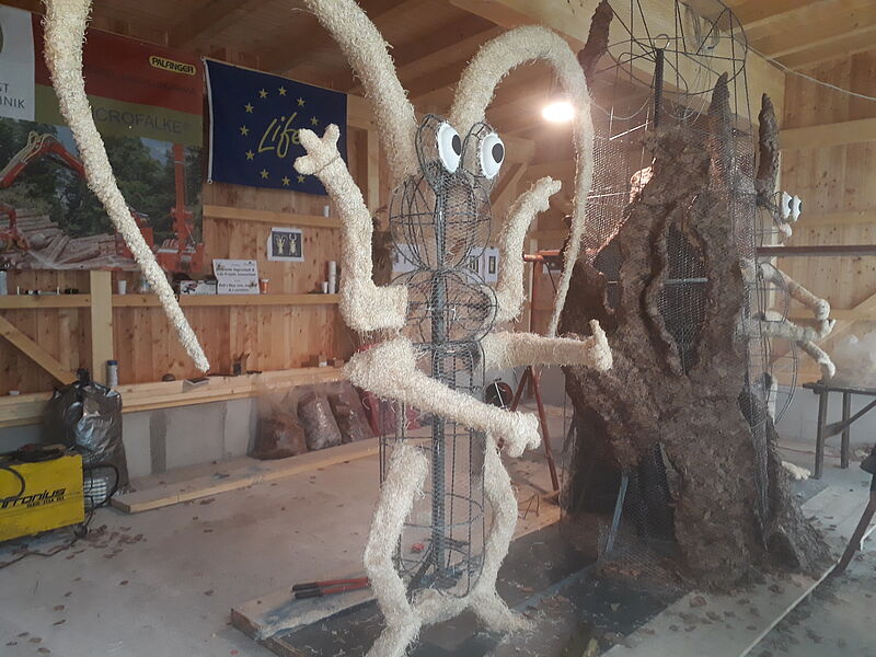 Ein zwei Meter hohes Gestell aus Draht in Form eines Alpenbockkäfers steht in einer Halle aus Holz.