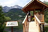 Eine kleine Hütte im Ausseerstil aus Holz wurde über dem Wasser auf Pfahlen errichtet. Eine Informationstafel steht davor. Ein Besucher wagt sich gerade die Rampe hinauf.