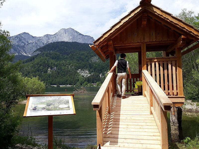 Eine kleine Hütte im Ausseerstil aus Holz wurde über dem Wasser auf Pfahlen errichtet. Eine Informationstafel steht davor. Ein Besucher wagt sich gerade die Rampe hinauf.