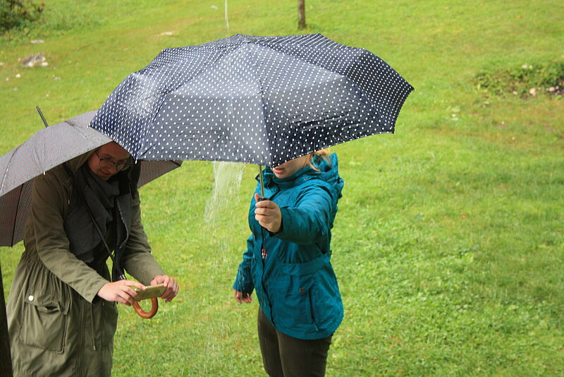 Zwei Mädchen stehen mit jeweils einem Schirm vor einer Regentonne, in die Wasser aus der Dachrinne läuft. Das eine Mädchen hta ein Handy und fimt die Situation , während das andere seinen Schirm so hält, dass der Wasserstrahl unterbrochen wird.