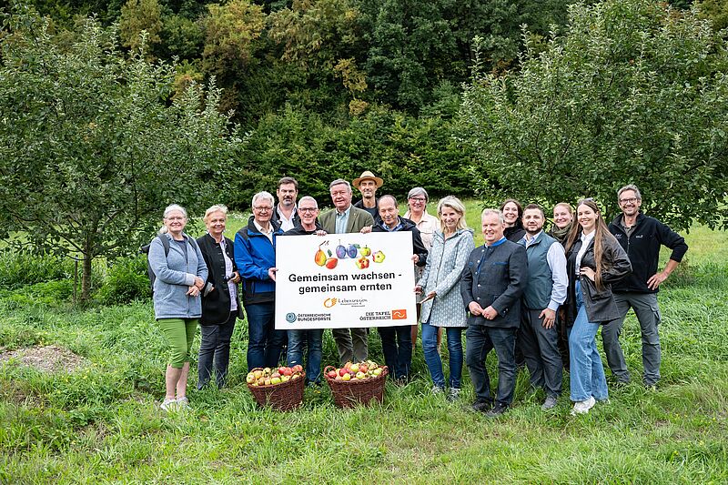 Vertreter von Biosphärenpark, Bundesforsten, Gemeinden und Tafel Österreich halten ein Schild auf dem "gemeinsam wachsen - gemeinsam ernten!" steht. Im Hintergrund ist eine Streuobstwiese.