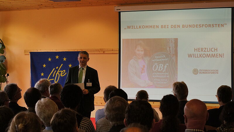 Ein Mann steht vor einem Publikum. Im Hintergrund sieht man die PowerPoint Präsentation und ein blaues Plakat mit der Aufschrift LIFE.