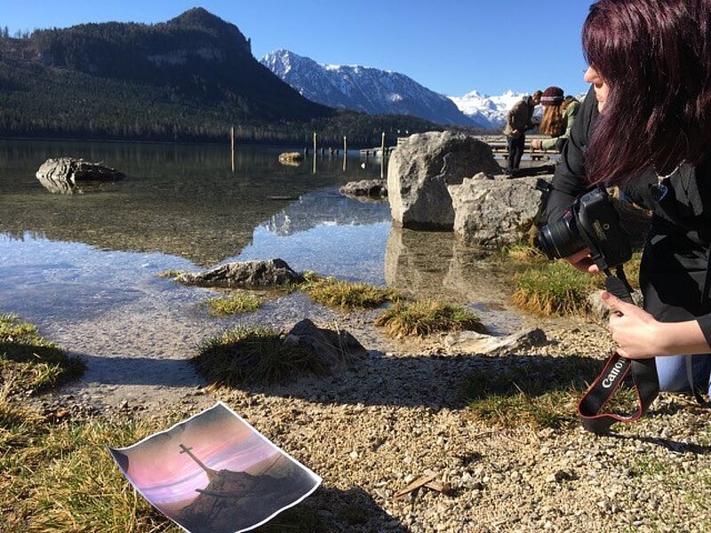 Am rechten Rand kniet eine Jugendliche mit einer Spiegelreflexkamera in der Hand. Sie schaut Richtung See. Links unten im Eck liegt ein ausgedrucktes A4 Blatt, auf dem ein Gipfelkreuz bei Sonnenuntergang zu sehen ist.