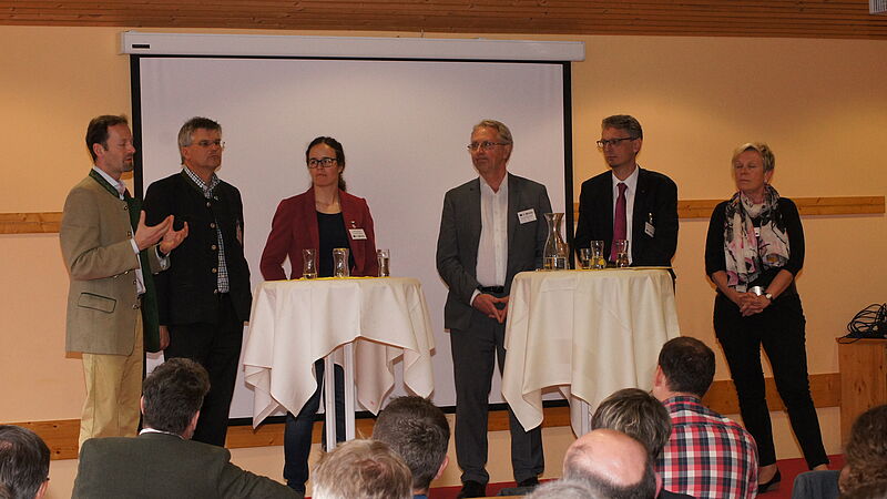 Sechs Personen, davon zwei Fraauen stehen auf einer Bühne hinter zwei Stehtischen. Der Mann ganz links erklärt gerade etwas und gestikuliert.