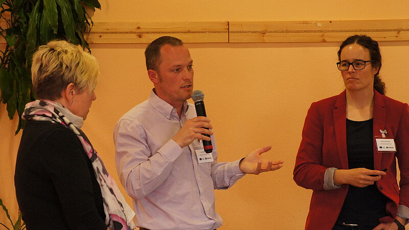 Ein Mann steht zwischen zwei Frauen und hält ein Mikrophon. Er antwortet mit einer Gestik auf eine Frage.